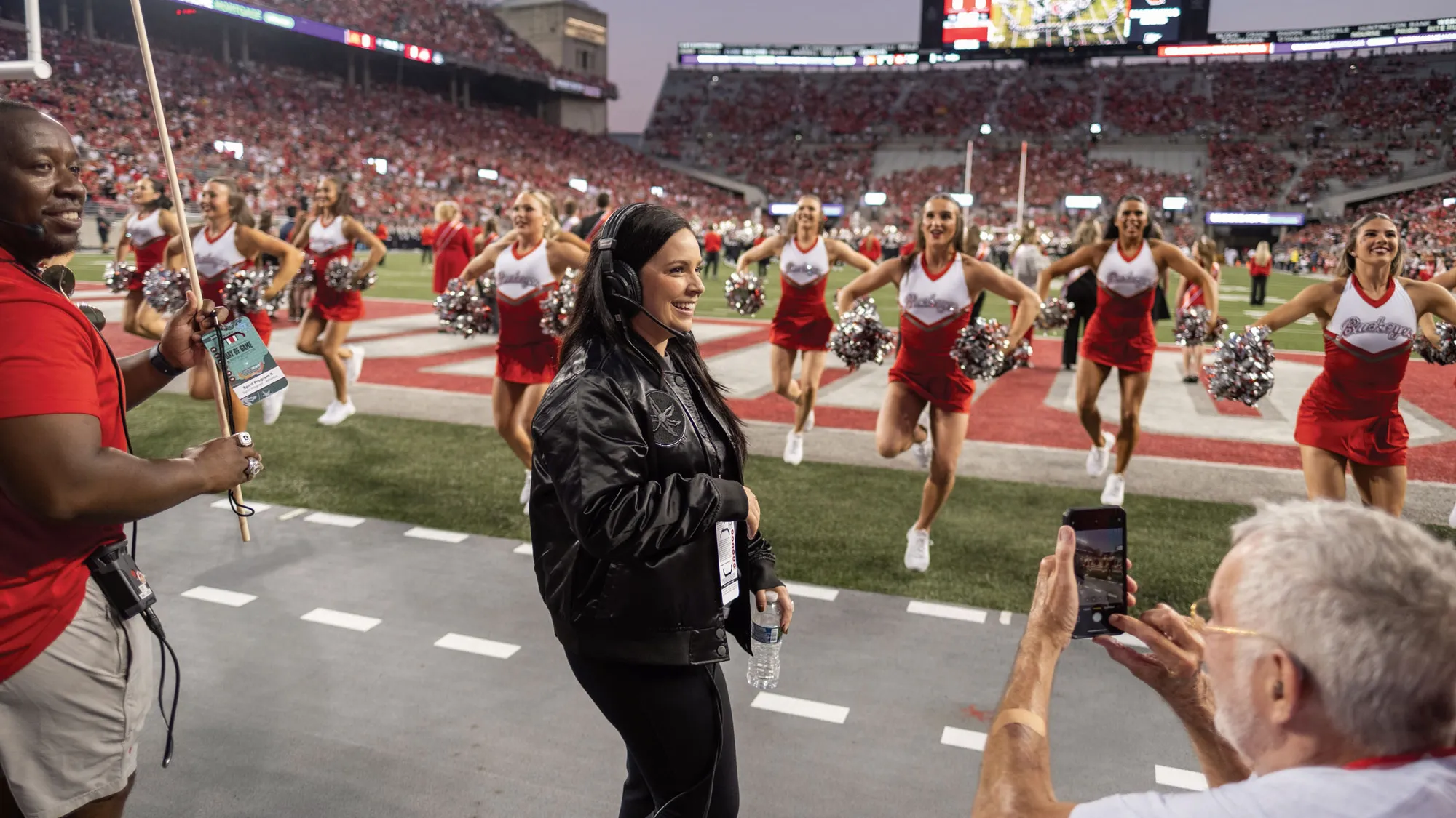 Melissa McGhee, a short white woman with long dark hair, smiles as she coaches the dance and cheer teams on the sidelines of an Ohio State football game. In Ohio Stadium, she wears a leather Ohio State coat and a headset.