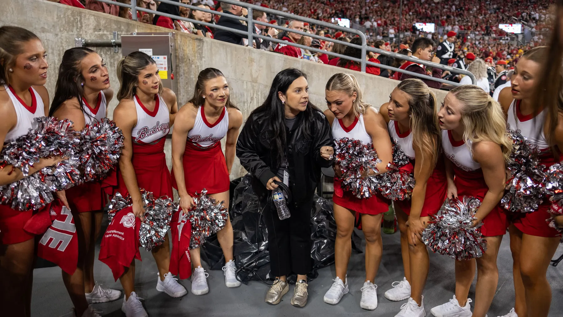 Standing arm in arm, the dance team bends forward to listen as Melissa talks. They're grouped in one of the ramps to the football field, and a sea of fans can be seen in the stands behind them.
