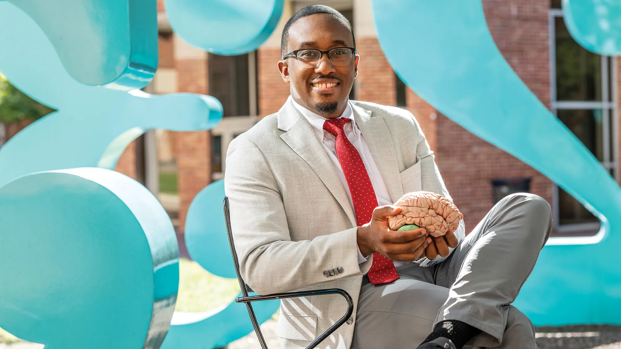 Jeremy Grant holds a model brain while sitting in a chair at the numbers sculptures on Ohio State's campus. He's a black man wearing a suit, tie and glasses. He has a friendly smile and his hair is short and neatly trimmed. 