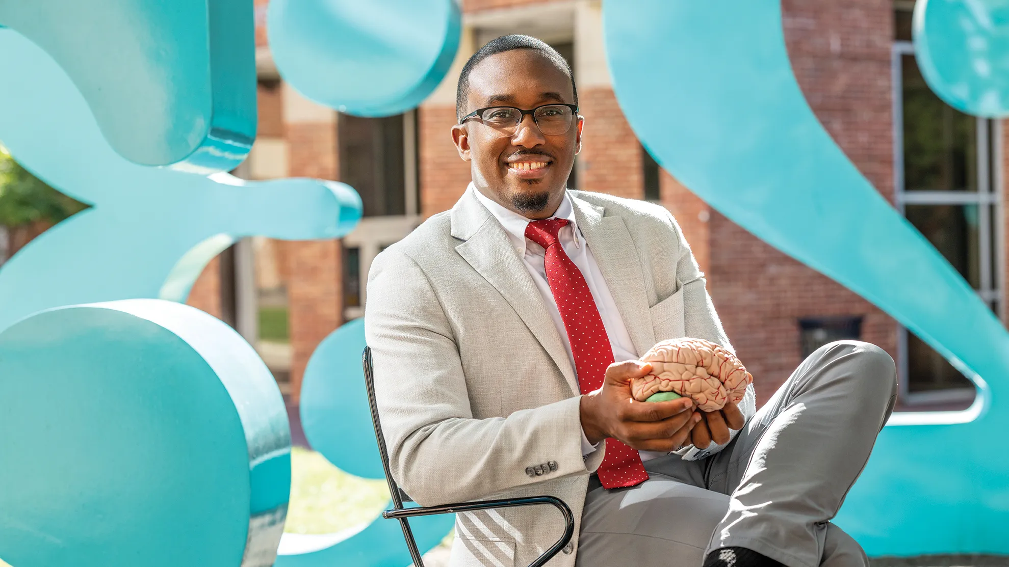 Jeremy Grant holds a model brain while sitting in a chair at the numbers sculptures on Ohio State's campus. He's a black man wearing a suit, tie and glasses. He has a friendly smile and his hair is short and neatly trimmed. 