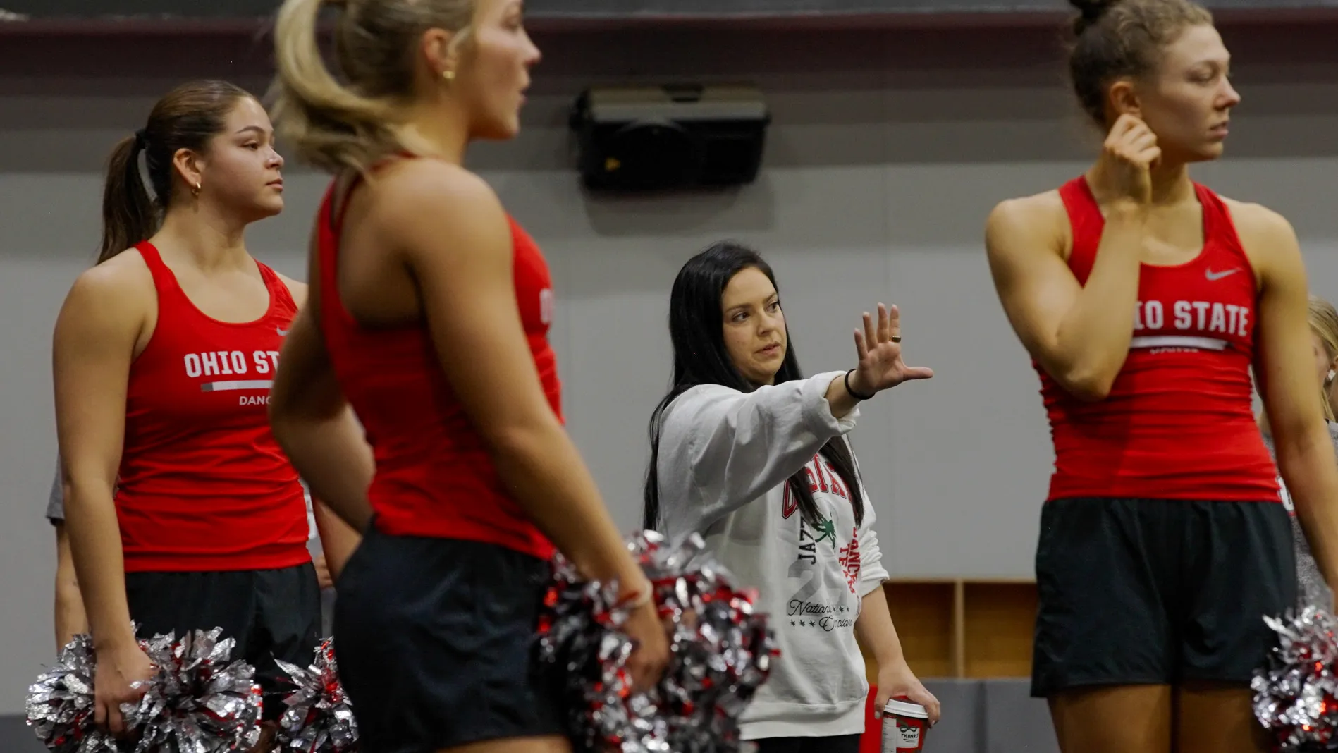 Melissa McGhee, a short, pretty white woman with long dark hair, coaches dance team members. She gestures with her hand reached out and they all look that way.
