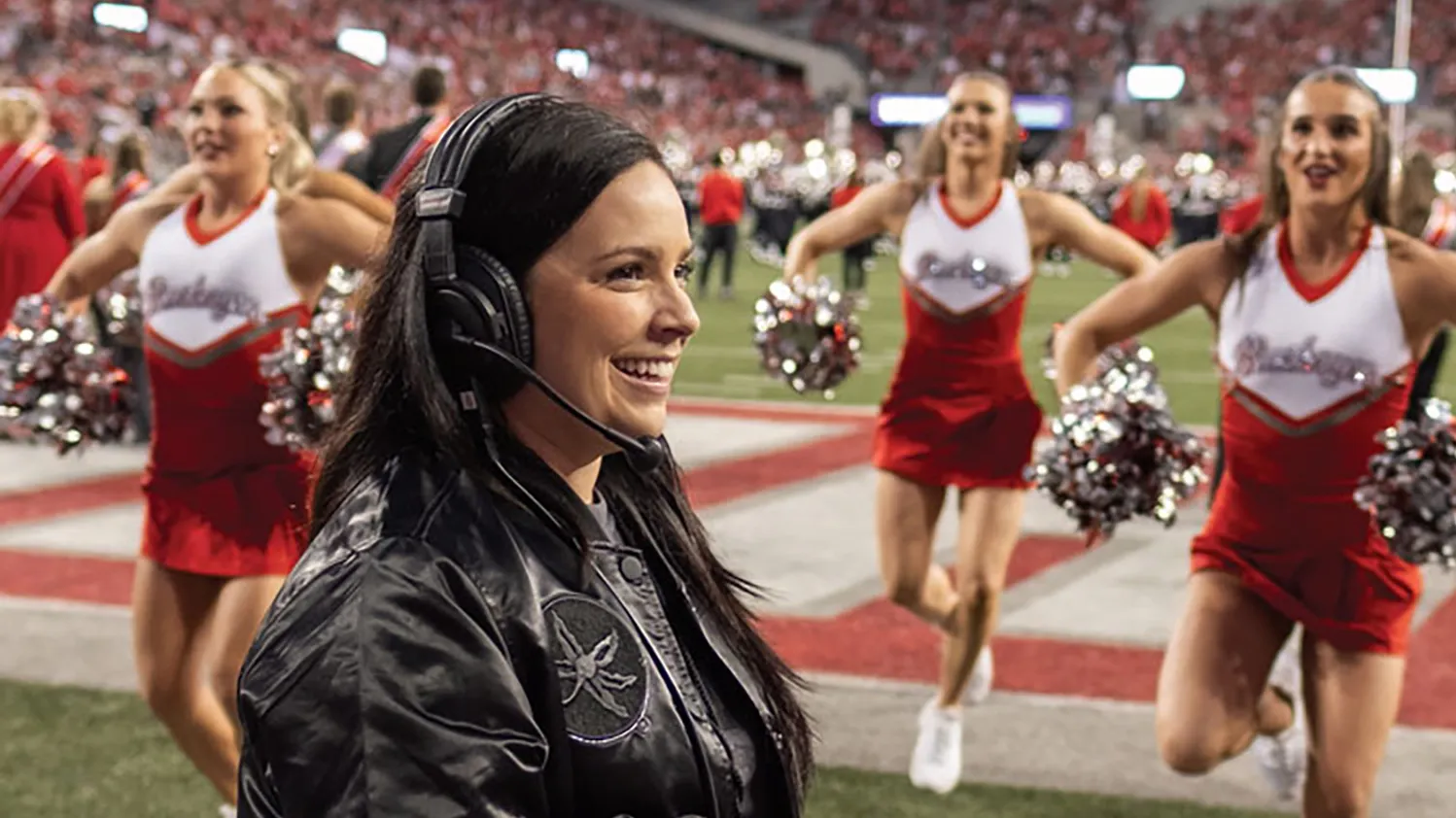 Melissa McGhee, a short white woman with long dark hair, smiles as she coaches the dance and cheer teams on the sidelines of an Ohio State football game. In Ohio Stadium, she wears a leather Ohio State coat and a headset.