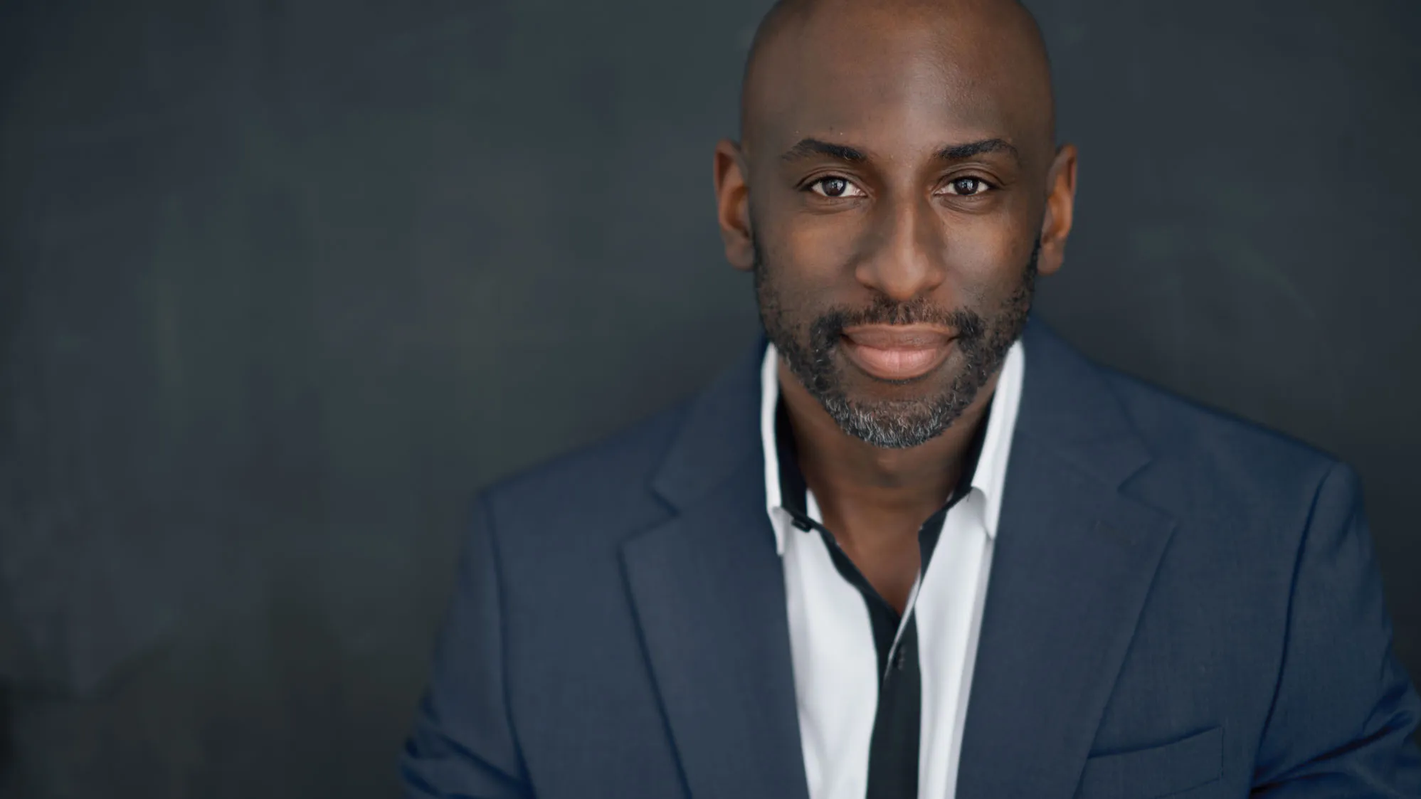 Rashad Chambers poses for a portrait wearing a suit and button down shirt. A young-ish black man, he appears stylish, calm, confident and proud.