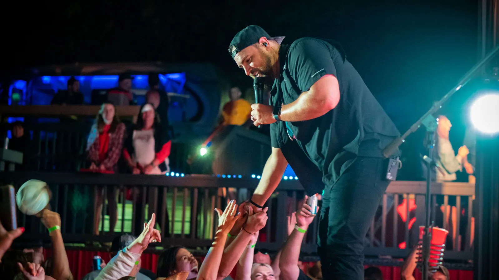A man holding a mic on stage reaches down to clasp hands with adoring audience members. He wears all black and a backwards baseball cap.