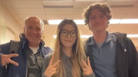 Three people grin as they pose for a casual, selfie-style photo. A grandpa and high school student stand with their TA between them.