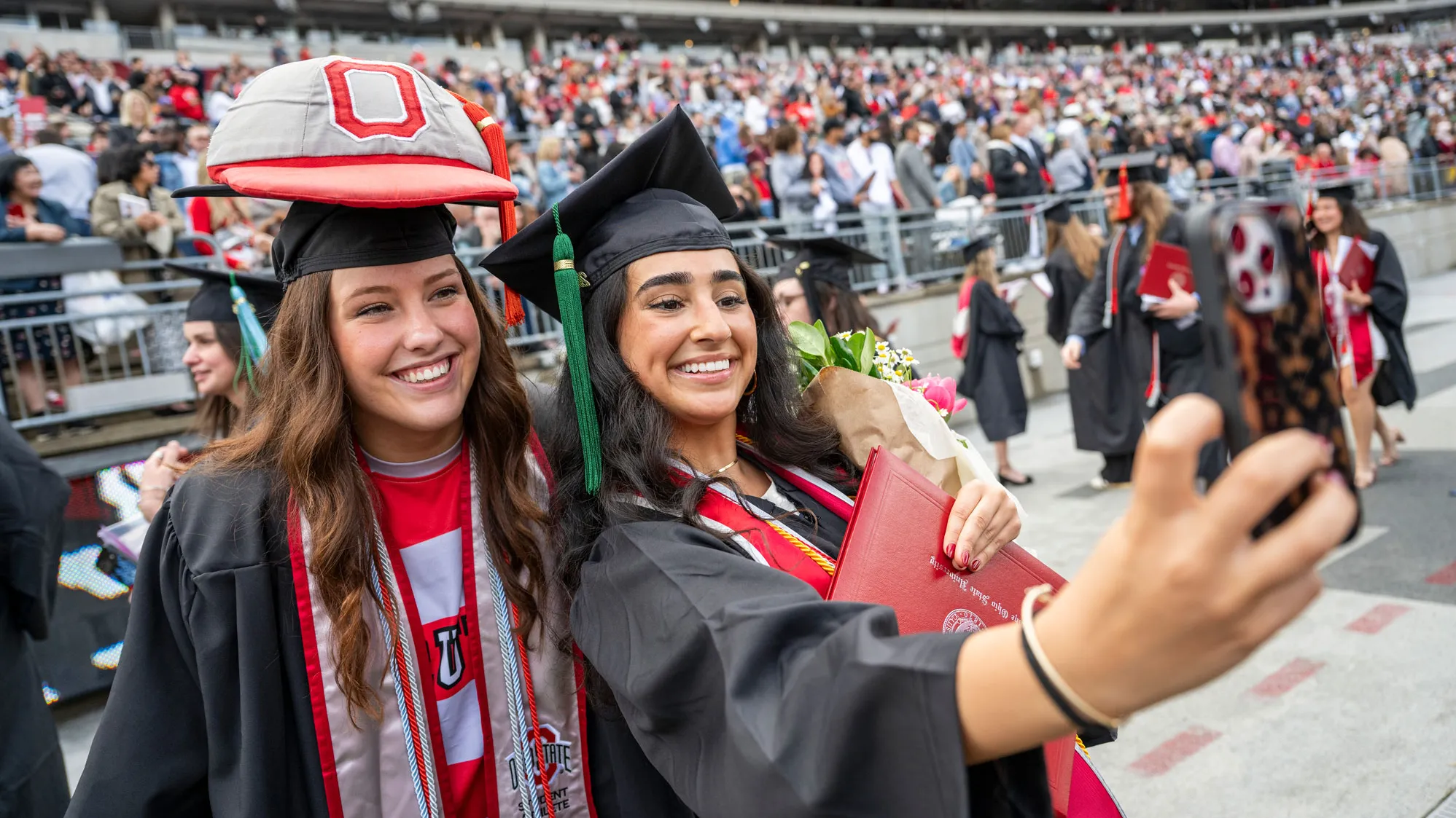 Two people in graduation caps and gowns take a selfie on a football stadium field, with one holding a diploma and bouquet and the other wearing Brutus’ giant cap on top of her graduation cap. A crowd of graduates and guests fills the stands in the background.