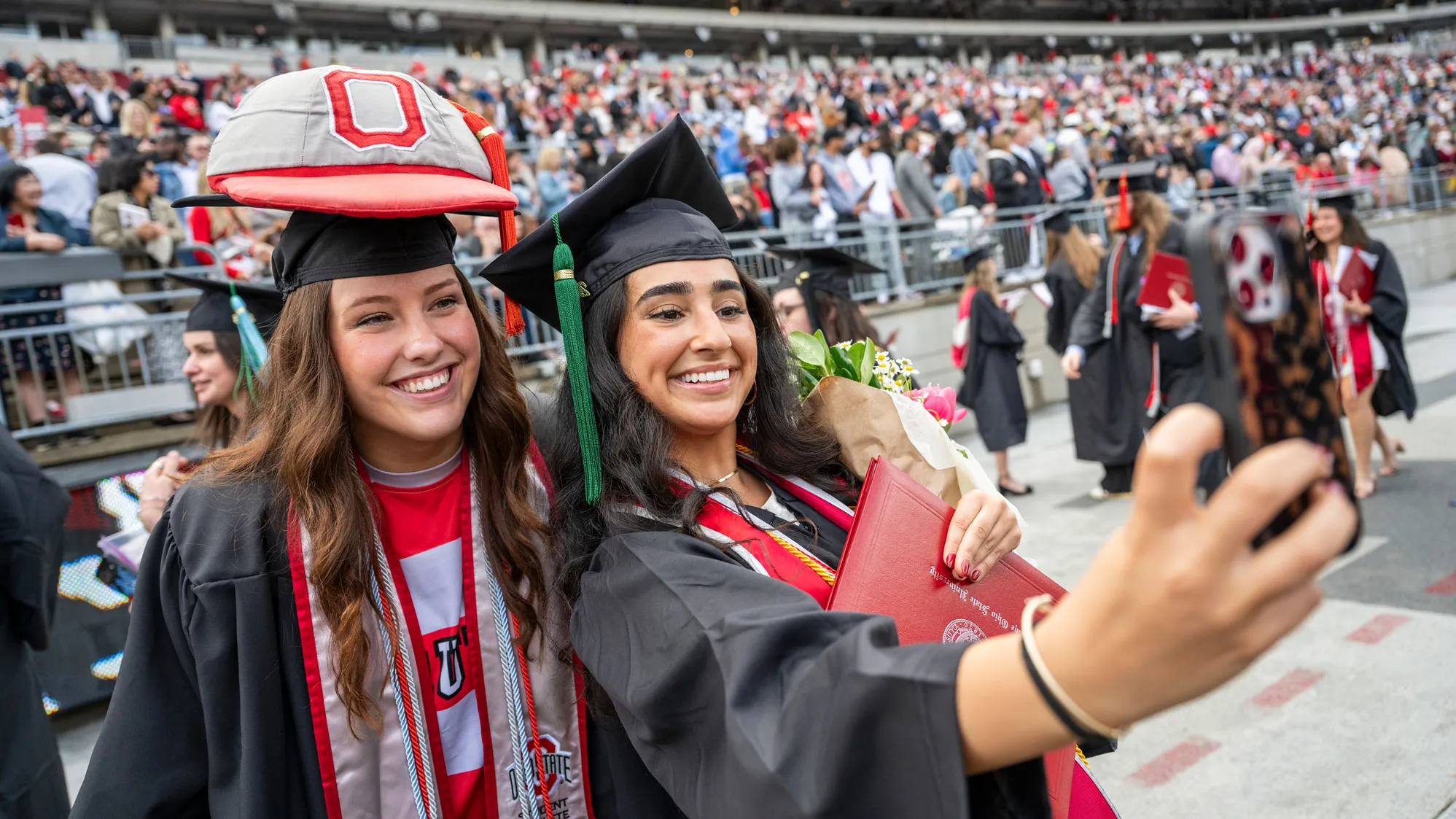 Two people in graduation caps and gowns take a selfie on a football stadium field, with one holding a diploma and bouquet and the other wearing Brutus’ giant cap on top of her graduation cap. A crowd of graduates and guests fills the stands in the background.