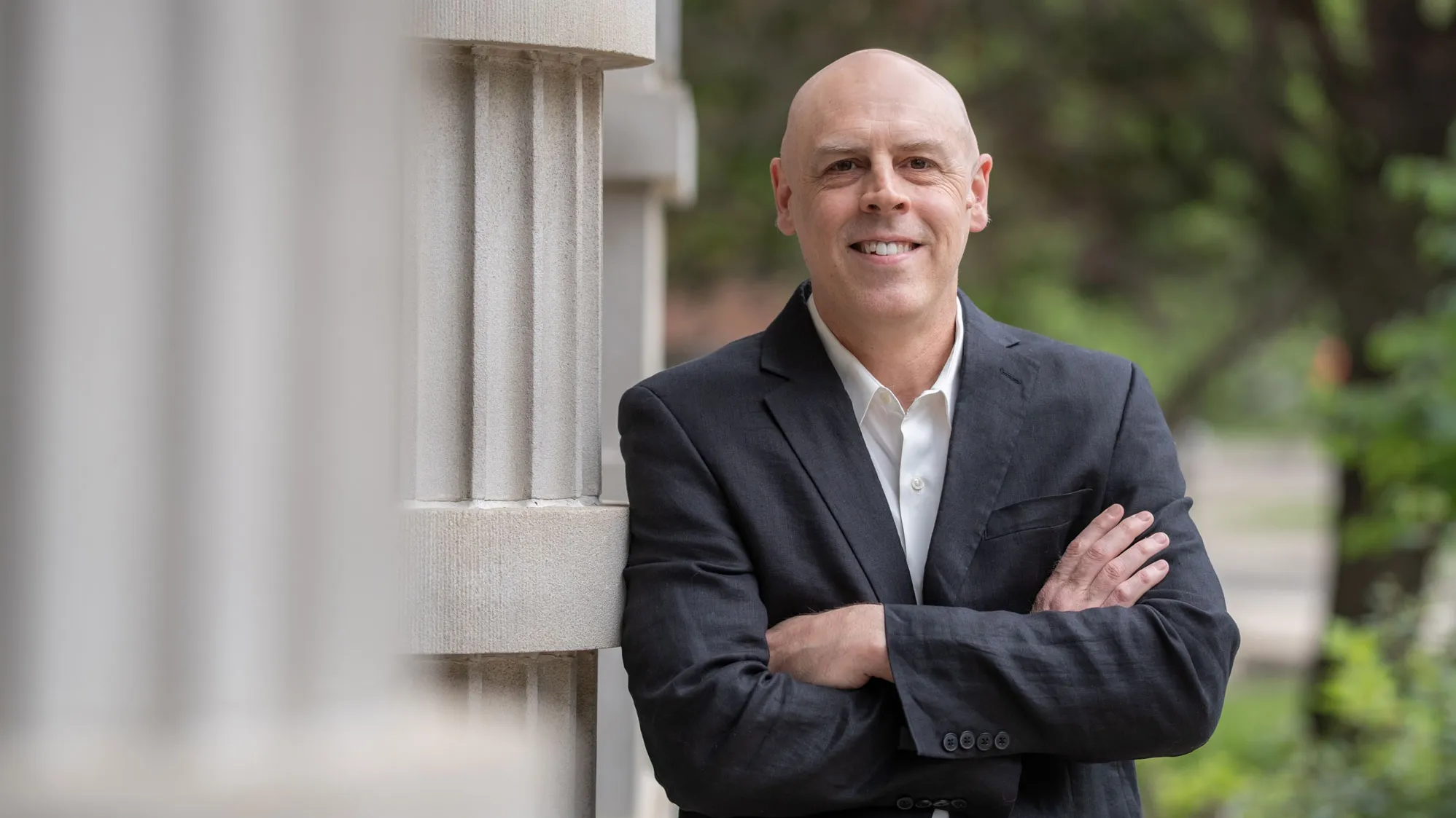 A professor smiles as he stands outdoors leaning against a column with arms crossed, wearing a collared shirt and suit jacket. He is a white man with a shaved head. Trees and a walkway appear in the background of the photo.