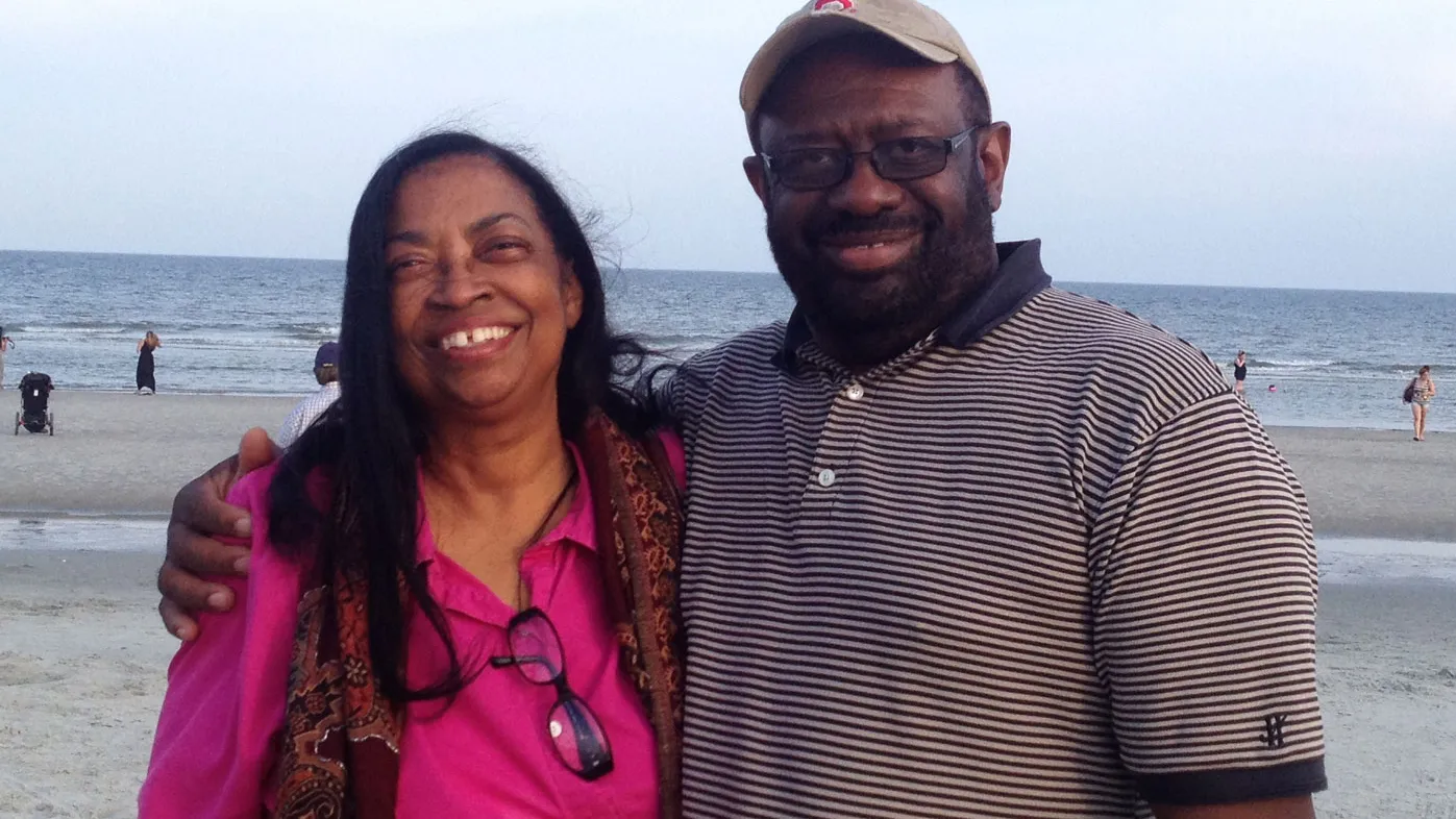 A husband and wife smile as they stand close together on a sandy beach with the ocean and a few distant beachgoers in the background.