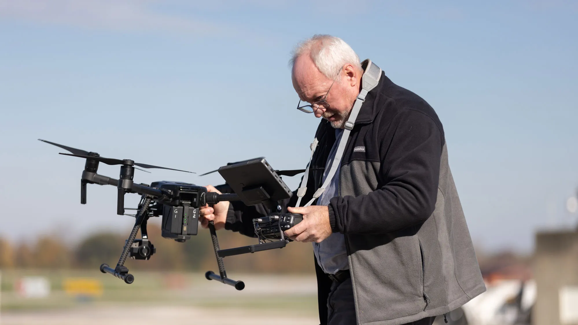 Outside at the airport, Dale stands outdoors holding a large drone with multiple rotors. He appears to be adjusting or inspecting the drone while also holding a controller mounted with a screen. It’s a sunny day, but cool enough that he’s wearing a jacket.