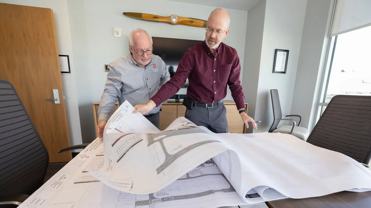 Two men stand at a conference table reviewing large architectural plans, with one person lifting a corner of the drawings. The setting is an office at Ohio State’s airport, with chairs around the table and a monitor and propeller blade mounted on the wall behind them.
