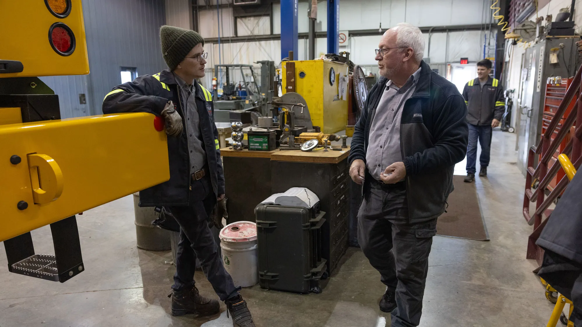 Two men talk inside a workshop filled with industrial equipment, tools, and machinery. The younger man wears a winter hat and leans against a large yellow vehicle used for runway maintenance. The other man, Dale, stands facing him. Workbenches, toolboxes, and various mechanical components are visible throughout the space, and another person is walking in the background.