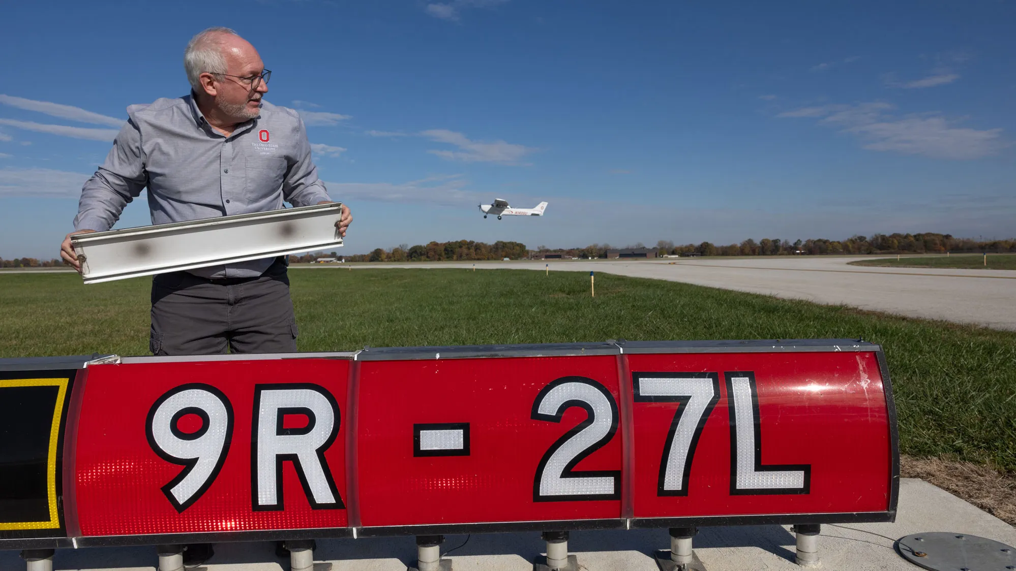 Dale stands behind a runway sign that reads “9R – 27L,” holding a long rectangular light fixture. He is beside the paved runway, and a small aircraft is visible in the air in the background under a clear sky.