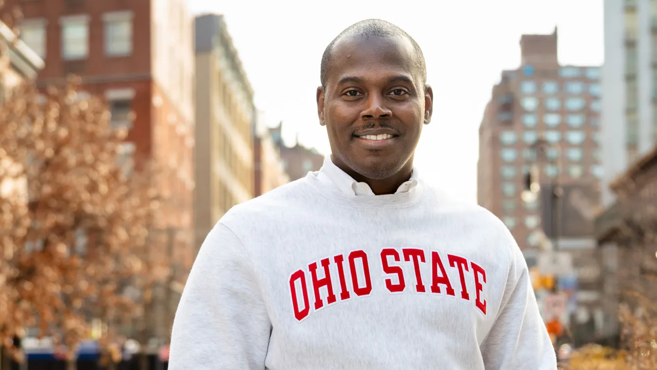 An African-American man smiles as he stands on a city street wearing a sweatshirt with the text “Ohio State” across the front, with thumbs tucked in his pockets and buildings and trees in the background. He looks like he's interested in what you have to say.