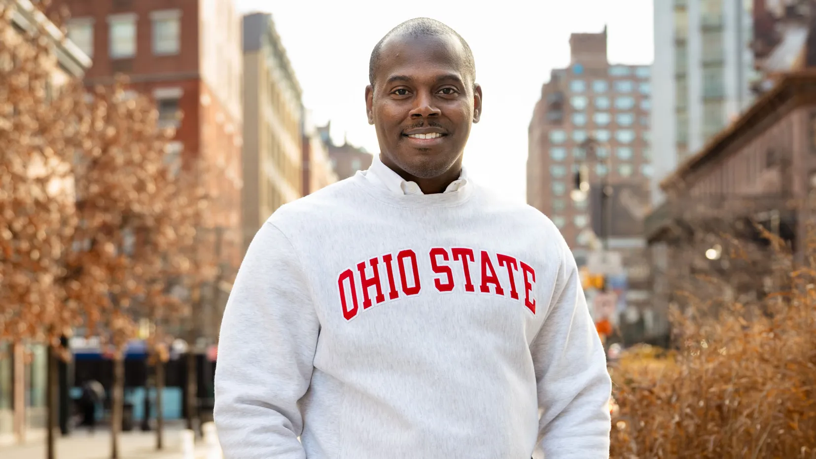 An African-American man smiles as he stands on a city street wearing a sweatshirt with the text “Ohio State” across the front, with thumbs tucked in his pockets and buildings and trees in the background. He looks like he's interested in what you have to say.