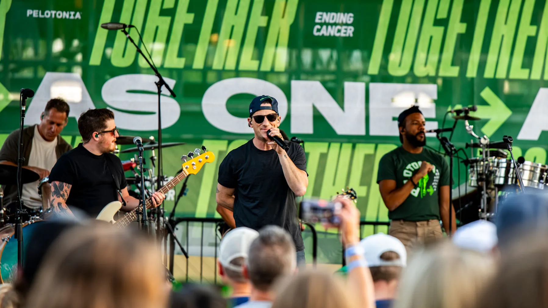 A band performs on an outdoor stage in front of a large backdrop with bold text including “TOGETHER” and “ENDING CANCER.” The musicians play instruments such as guitar, bass, drums, and percussion, while the main singer stands at the front holding a microphone. An audience dances and cheers in front of them.