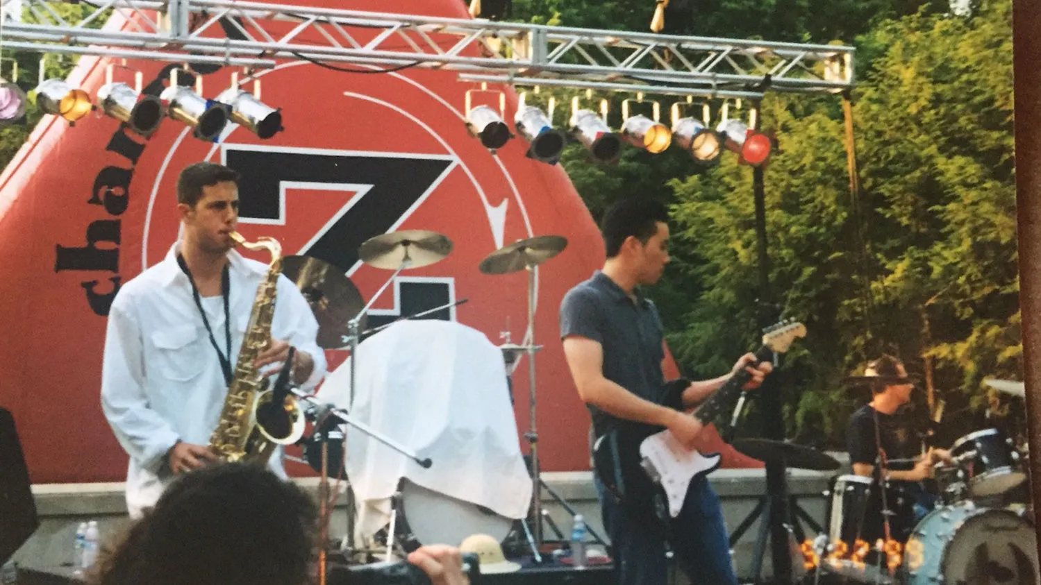 At a small Columbus festival, several musicians perform on an outdoor stage. One plays a saxophone, another plays an electric guitar, and a third plays a drum set. Trees fill the background.