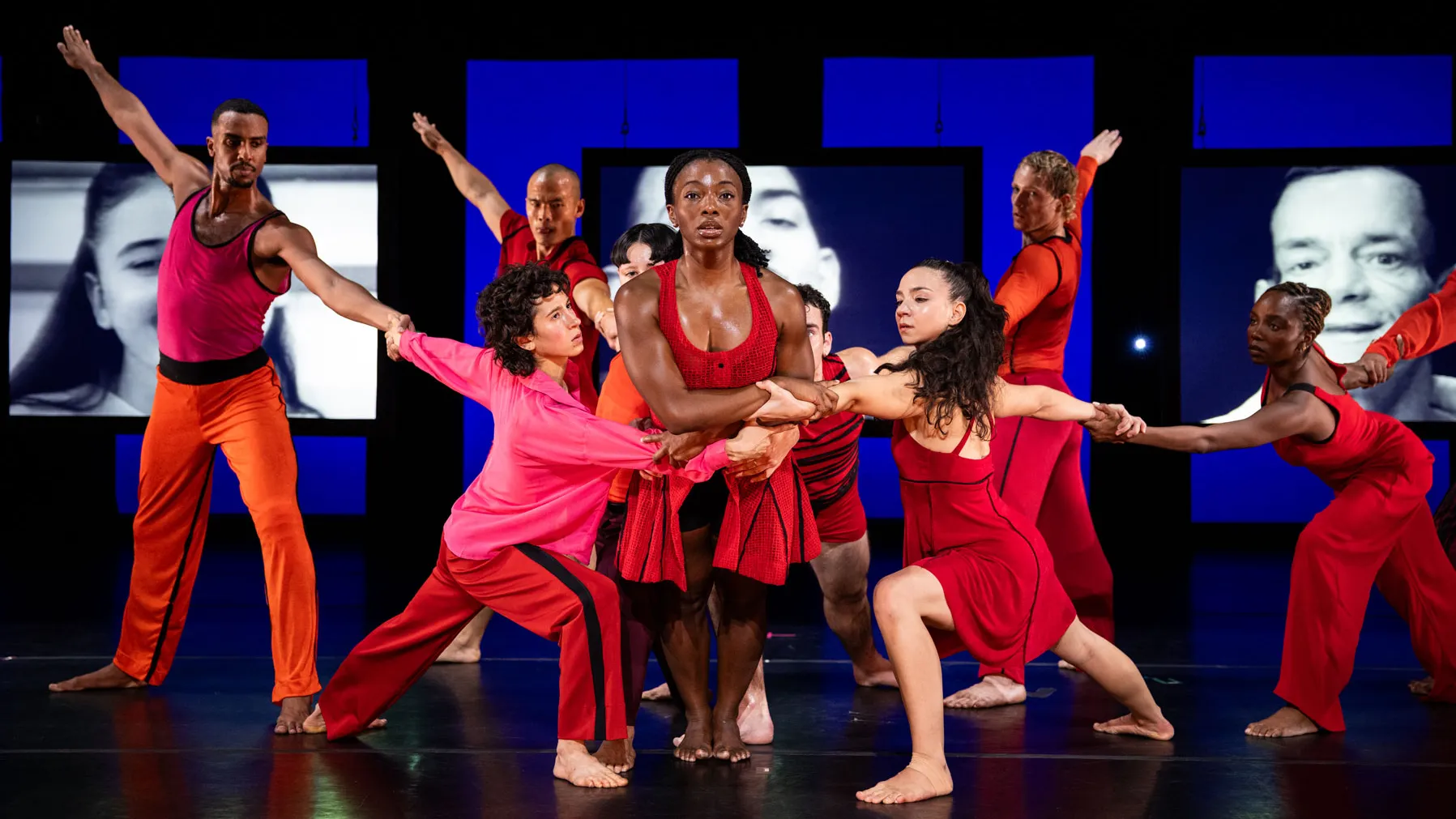 A group of dancers onstage perform a synchronized pose, holding onto one another in an interconnected formation. They wear brightly colored costumes. Behind them, large screens display black and white images, adding contrast to the vivid lighting and dynamic movement onstage.
