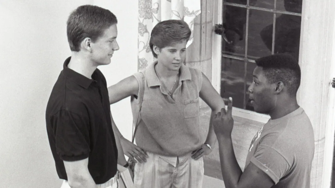 On a staircase landing, three college students engage in conversation. A large window with patterned curtains is behind them, and the wooden staircase features turned balusters and a decorative newel post. Of the three, one is woman and two are men; one is Black and two are white. They’re dressed in styles common in the 1980s.