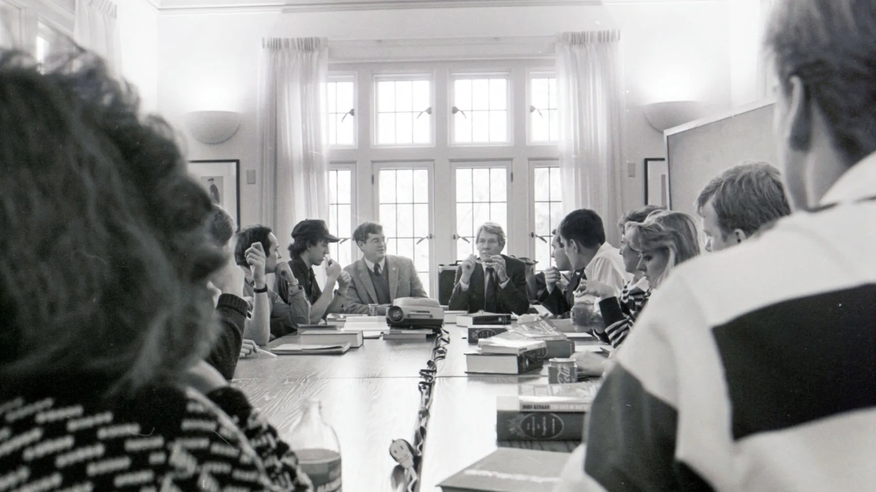 A group of college students engage in a serious discussion as they sit around a long table in a bright room with multi paned windows. Books, papers, and a few bottles are spread across the table. A couple of people at the far end appear to be faculty or staff.