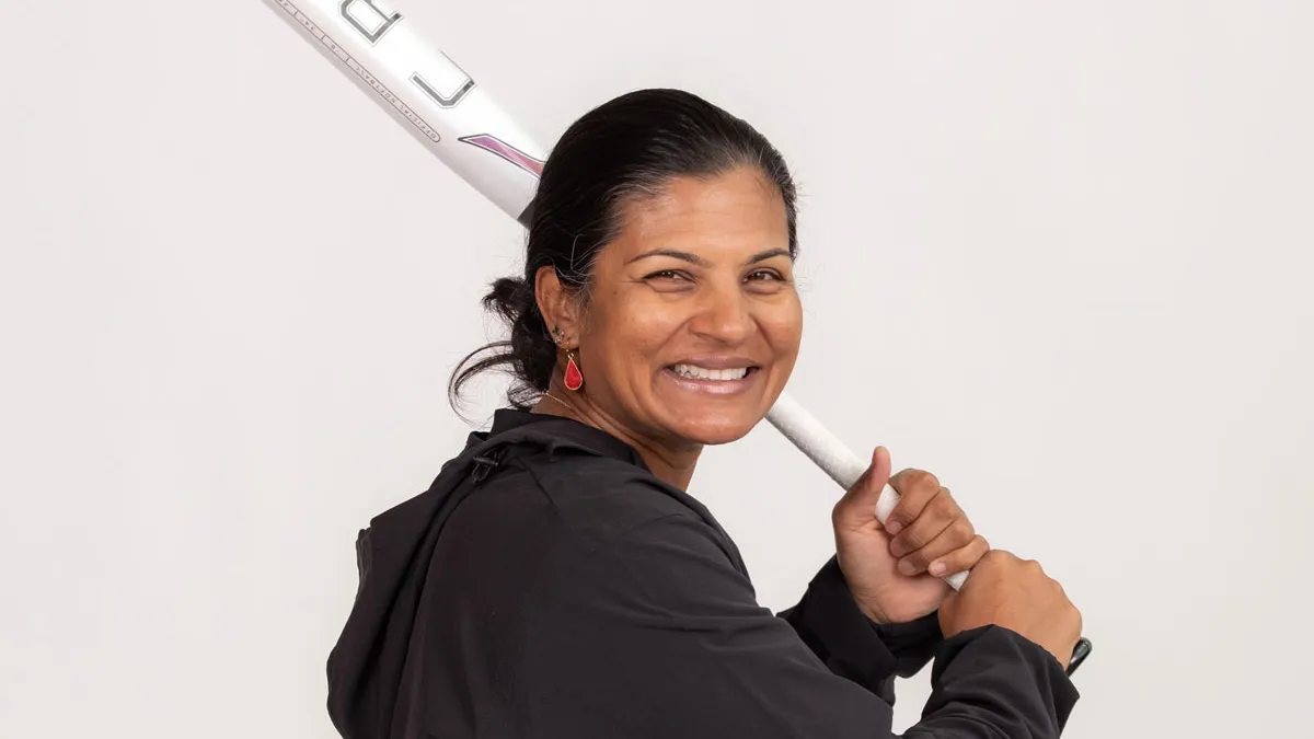 Kirin Kumar giggles as she poses in a batting stance, holding a baseball bat over one shoulder as if she’s getting ready for a pitch. 