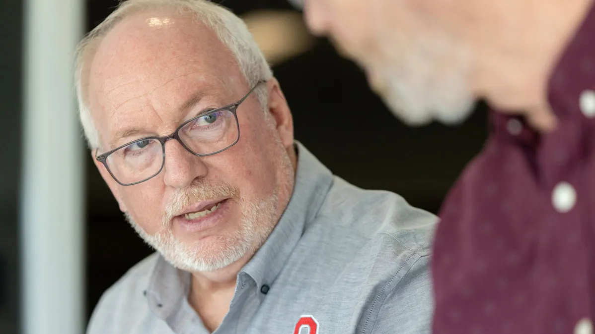 Dale Gelter talks to a colleague with a serious expression. He’s an older man with neat white hair and glasses and wears a button-down shirt with an embroidered Block O.