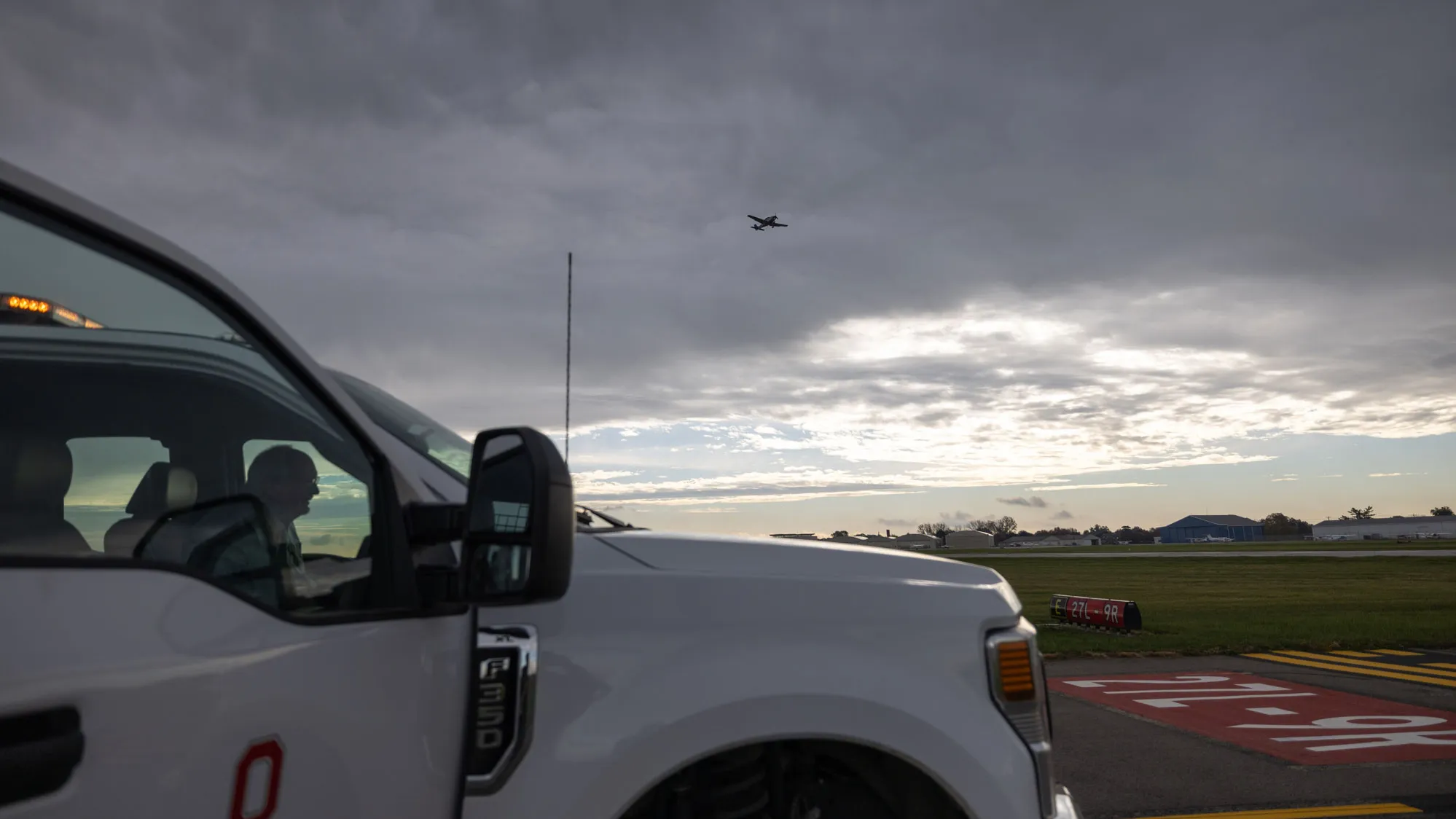 A person sits inside a parked truck near an airport runway, with part of the person's face visible through the window. A plane flies overhead beneath a cloudy sky.