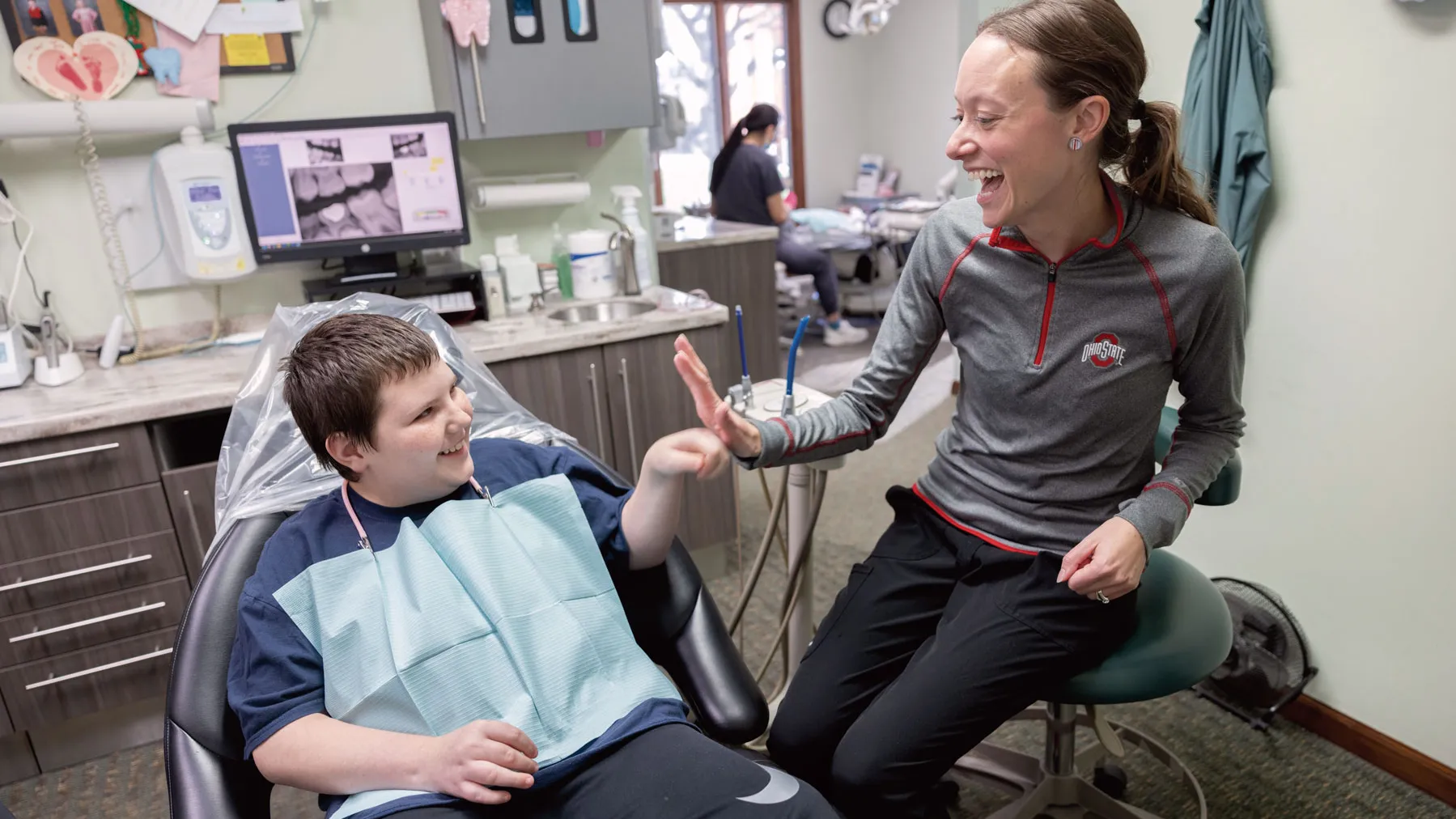 A dentist gives a high five to a young patient's fist bump. He sits in a chair with the paper bib people wear to be examined. And she wears an Ohio State shirt. They're both grinning as the watch each other.