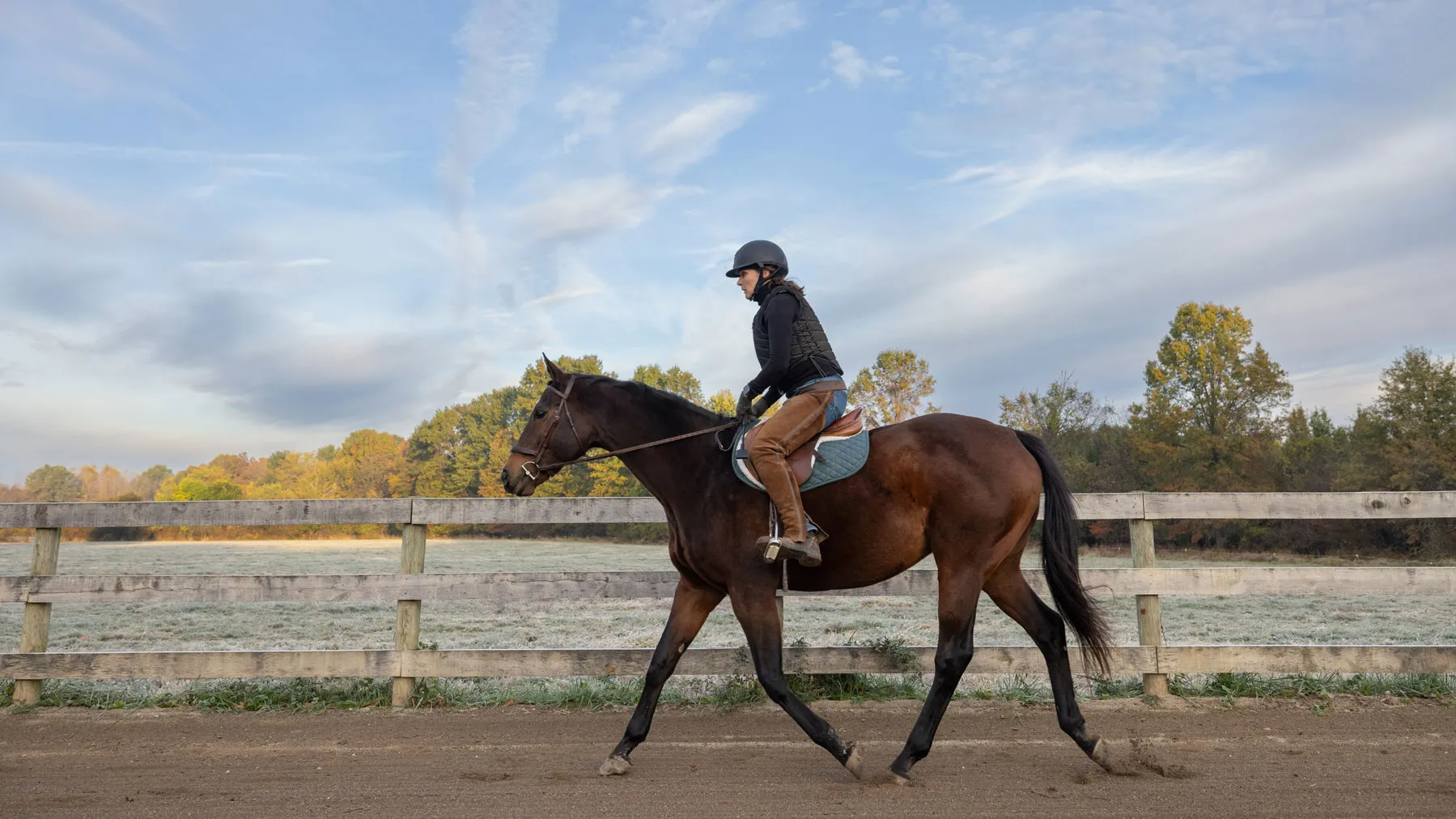 Lloyd wears a helmet and protective riding gear while riding a horse a fenced dirt path, with open fields and trees in the background under a wide sky.