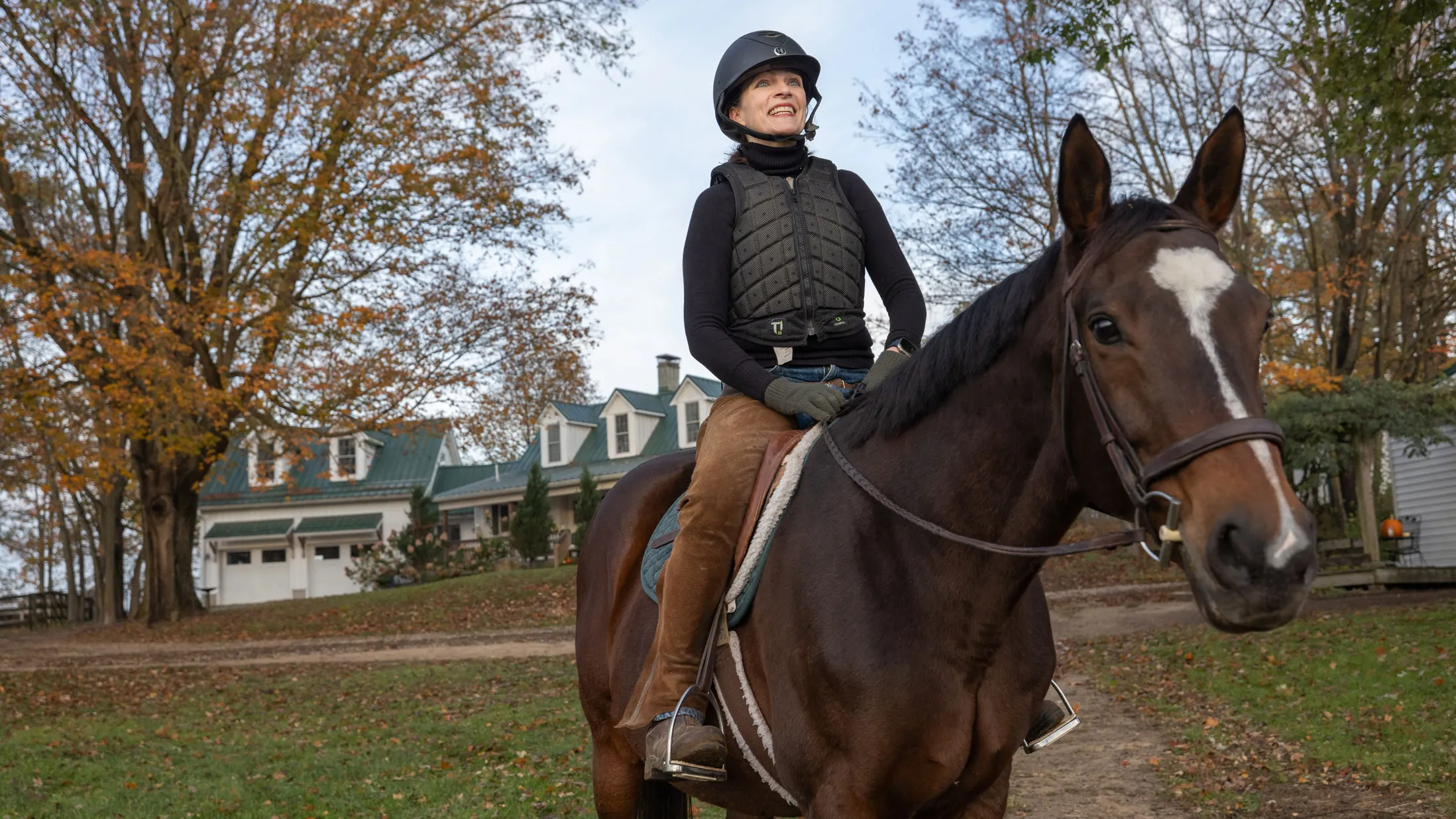 Kathy Lloyd smiles as she sits on a horse outside her riding stables on a crisp autumn day. She wears a helmet and protective riding vest sits and the horse looks directly at the camera as she looks off into the distance.