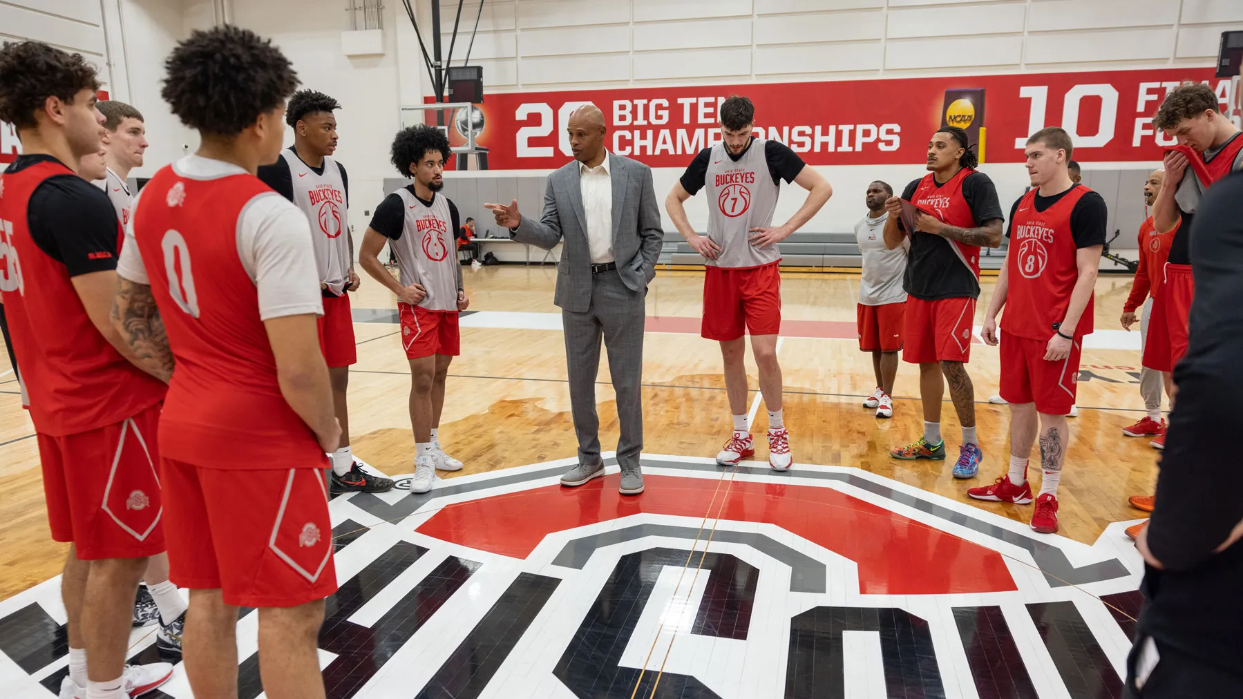 Ohio State men’s basketball players wear practice uniforms and stand in a circle at midcourt in a gym, listening to Kellogg as he talks to them. He wears a suit.