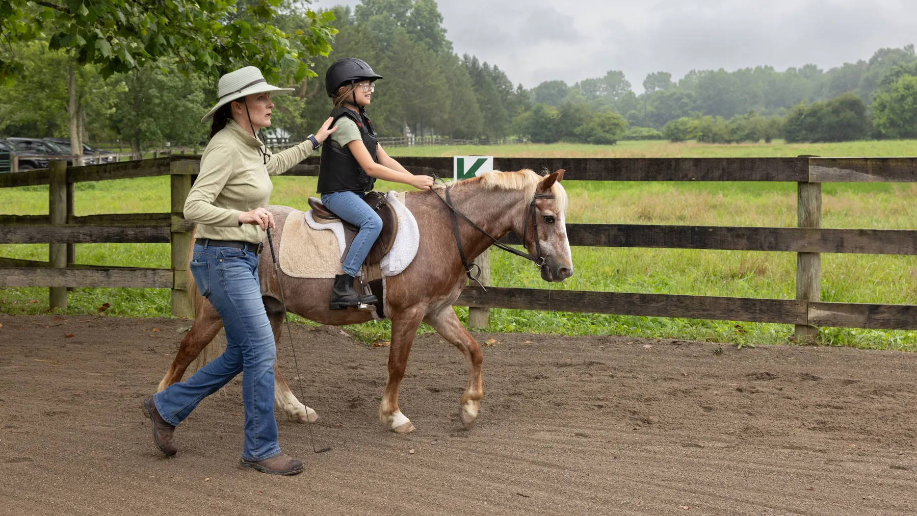 Lloyd walks alongside a small horse while guiding a young rider who is seated in the saddle. They’re next to an outdoor riding area bordered by a wooden fence, with fields and trees in the background.