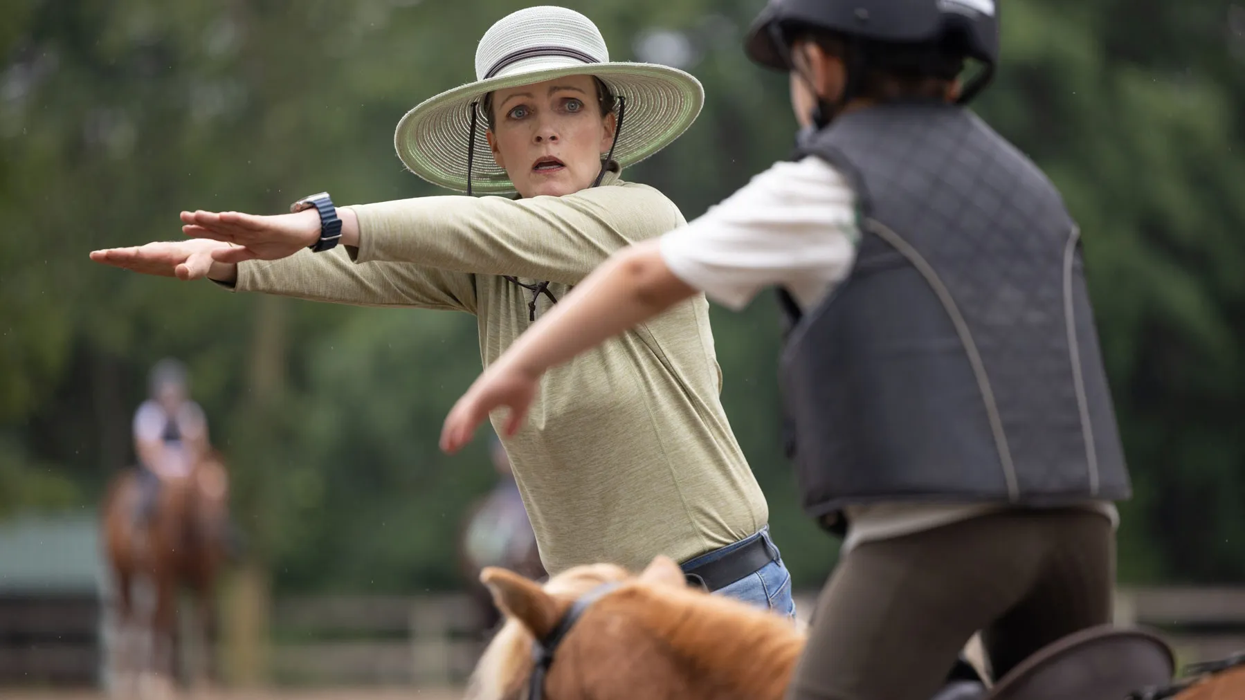 Lloyd demonstrates an arm stretching motion while a child rider on a horse mirrors her movement during an outdoor lesson.