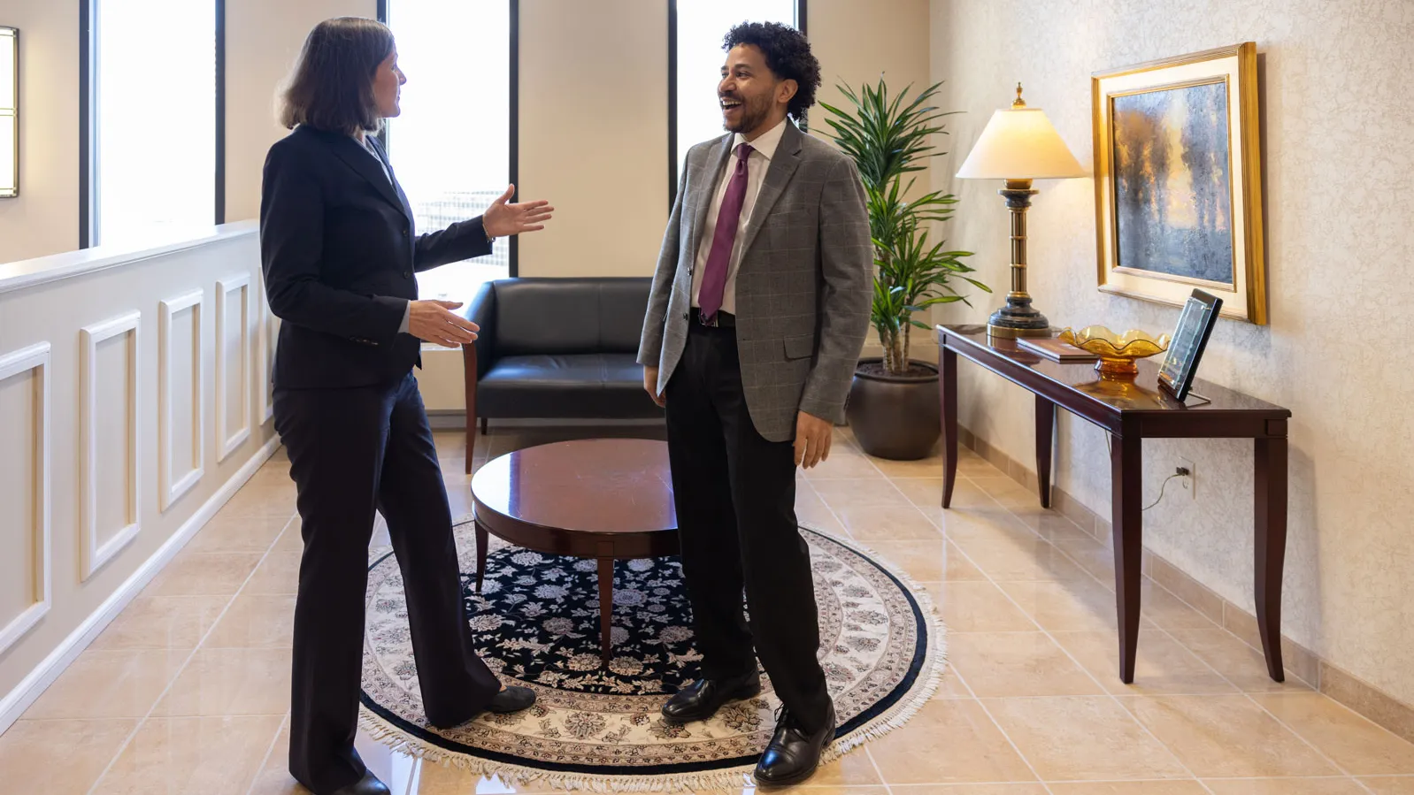 In a lobby area, Lloyd laughs as she talks with a coworker, a man in a suit with curly hair, during a conversation. Nearby are a round table, with seating, a console table, and framed artwork.