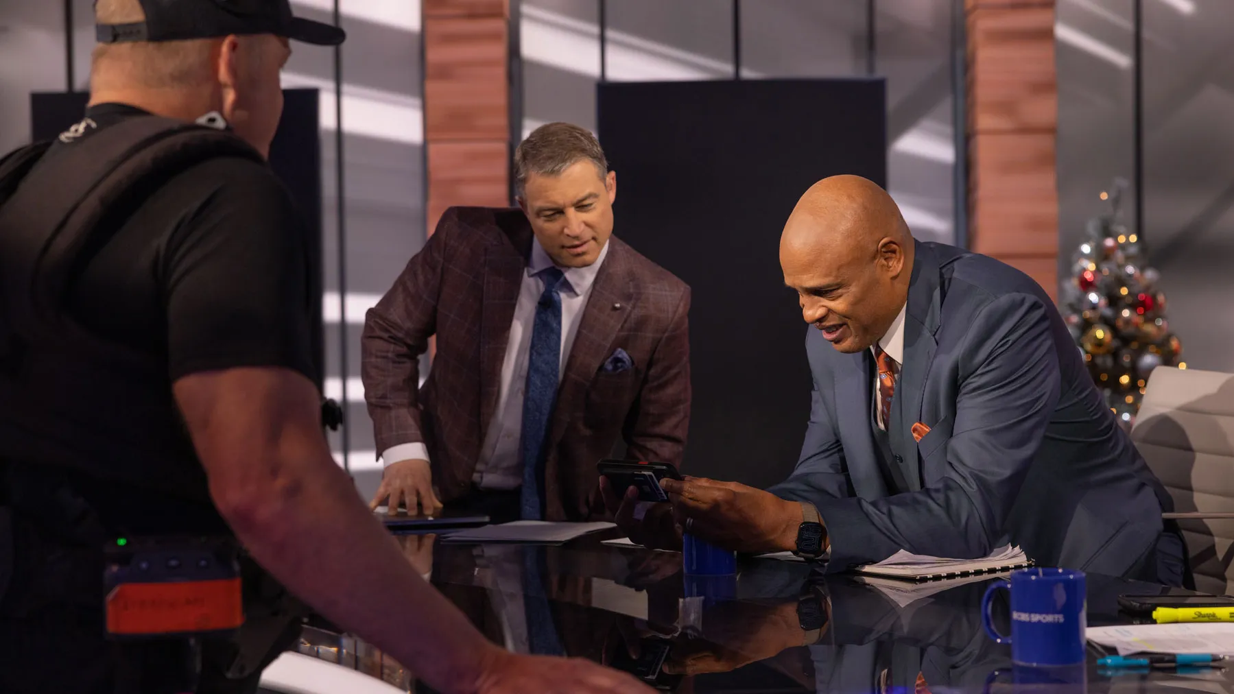 Kellogg and host Adam Zucker laugh as they lean over Kellogg’s phone at the studio news desk, while another man wearing production gear stands across the desk from them watching, too. Papers, cups, and studio equipment are on the desk, with set lighting visible in the background.