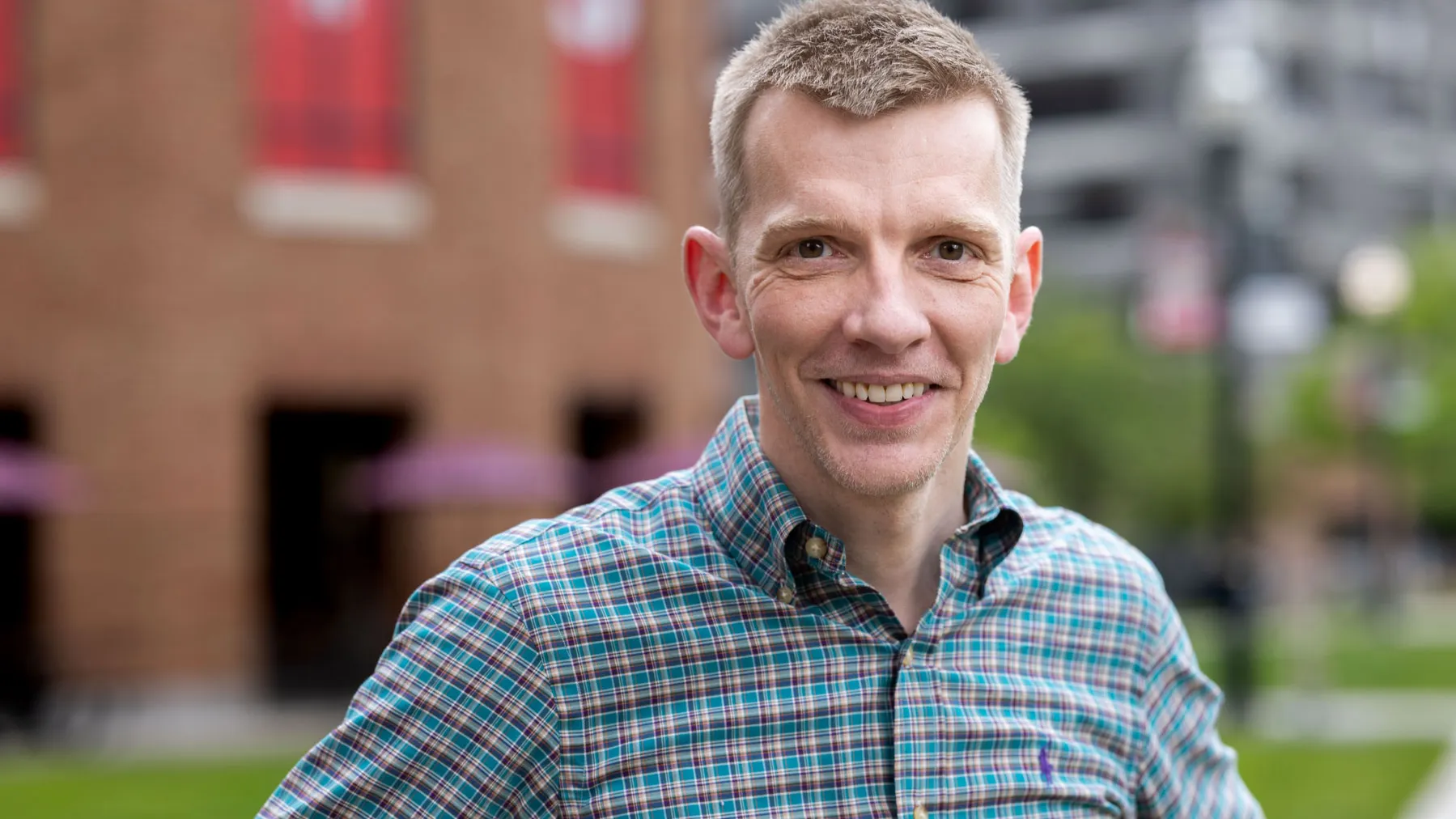 Angus Fletcher, a white man with neatly trimmed short hair, smiles and wears a patterned button-up shirt while standing outdoors with hands on hips. A brick building and a campus background with greenery appear behind him.