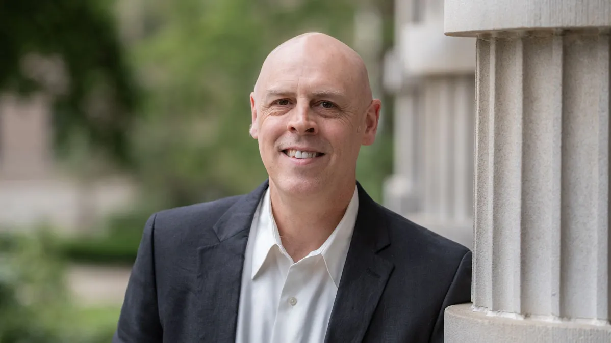 A professor smiles as he stands outdoors leaning against a column with arms crossed, wearing a collared shirt and suit jacket. He is a white man with a shaved head. Trees and a walkway appear in the background of the photo.