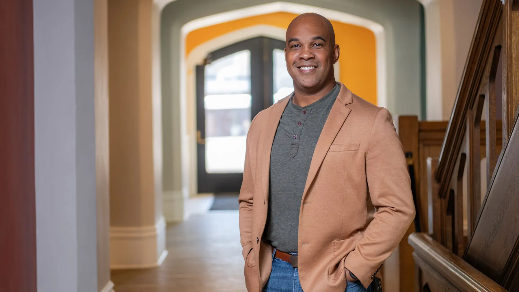 A Black man smiles as he stands in a hallway with hands in pockets, wearing a sport jacket and jeans, positioned near a wooden staircase with an arched doorway visible in the background. 