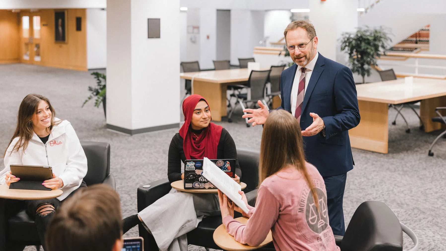 A group of law students sit together in a lounge-style area with small tables, using laptops and tablets, while Professor Dennis Hirsch stands and speaks with them. He’s talking with his hands as he explains a point.