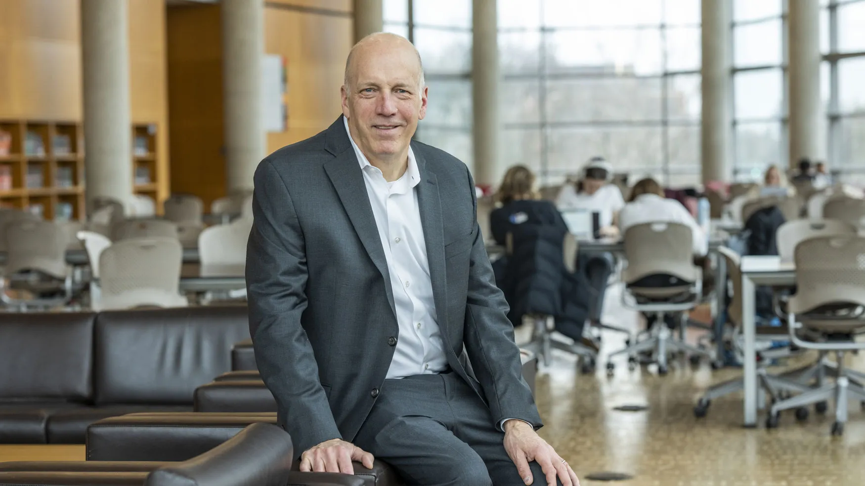 An older white man wearing a suit smiles as he sits on the edge of an upholstered chair in a spacious study room inside Thompson Library. Groups of people work at tables in the background and a full wall of windows and columns curves around the far side of the room.