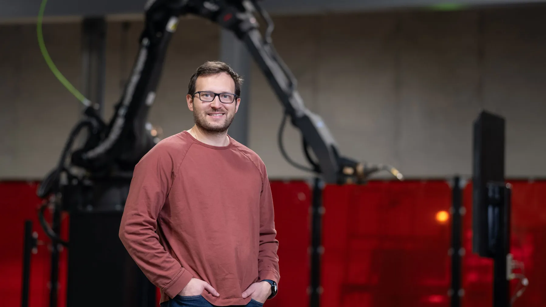 A white man smiles and stands inside a factory with his hands in pockets. Behind him is a giant robotic arm. He wears a sweatshirt, glasses and has neat hair and beard.