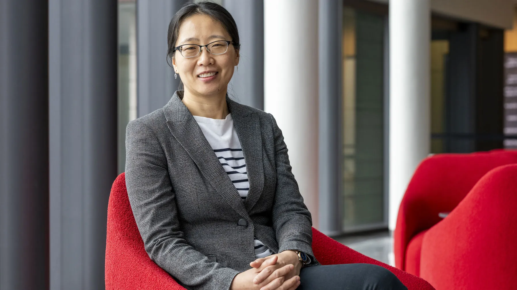 An Asian woman smiles as she sits in a modern indoor space on a curved chair, wearing a fitted blazer over a striped shirt with hands resting together. Tall windows, stately columns and additional chairs are visible in the background.