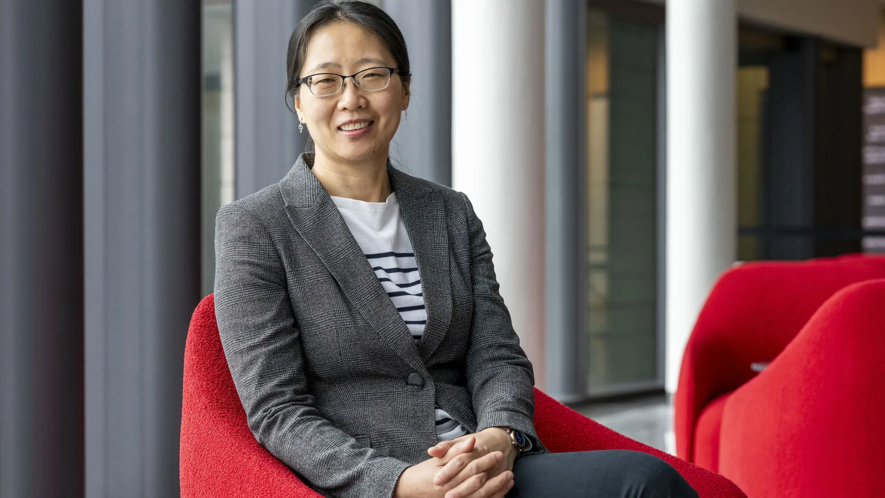 An Asian woman smiles as she sits in a modern indoor space on a curved chair, wearing a fitted blazer over a striped shirt with hands resting together. Tall windows, stately columns and additional chairs are visible in the background.