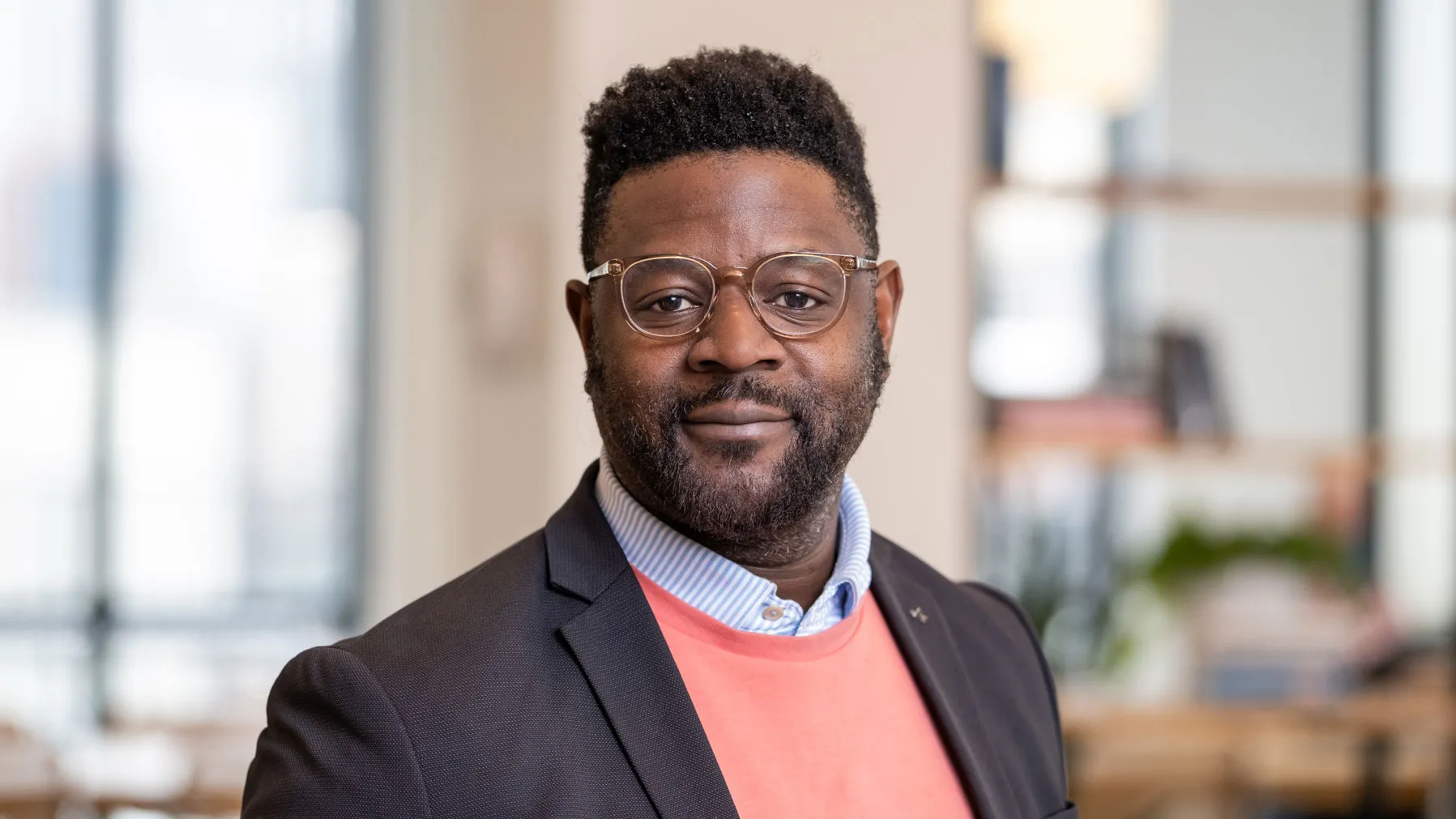 A Black man slightly smiles as he poses for this portrait in a modern office setting with shelves and large windows in the background. He wears glasses and sport coat over over a collared shirt and sweater.