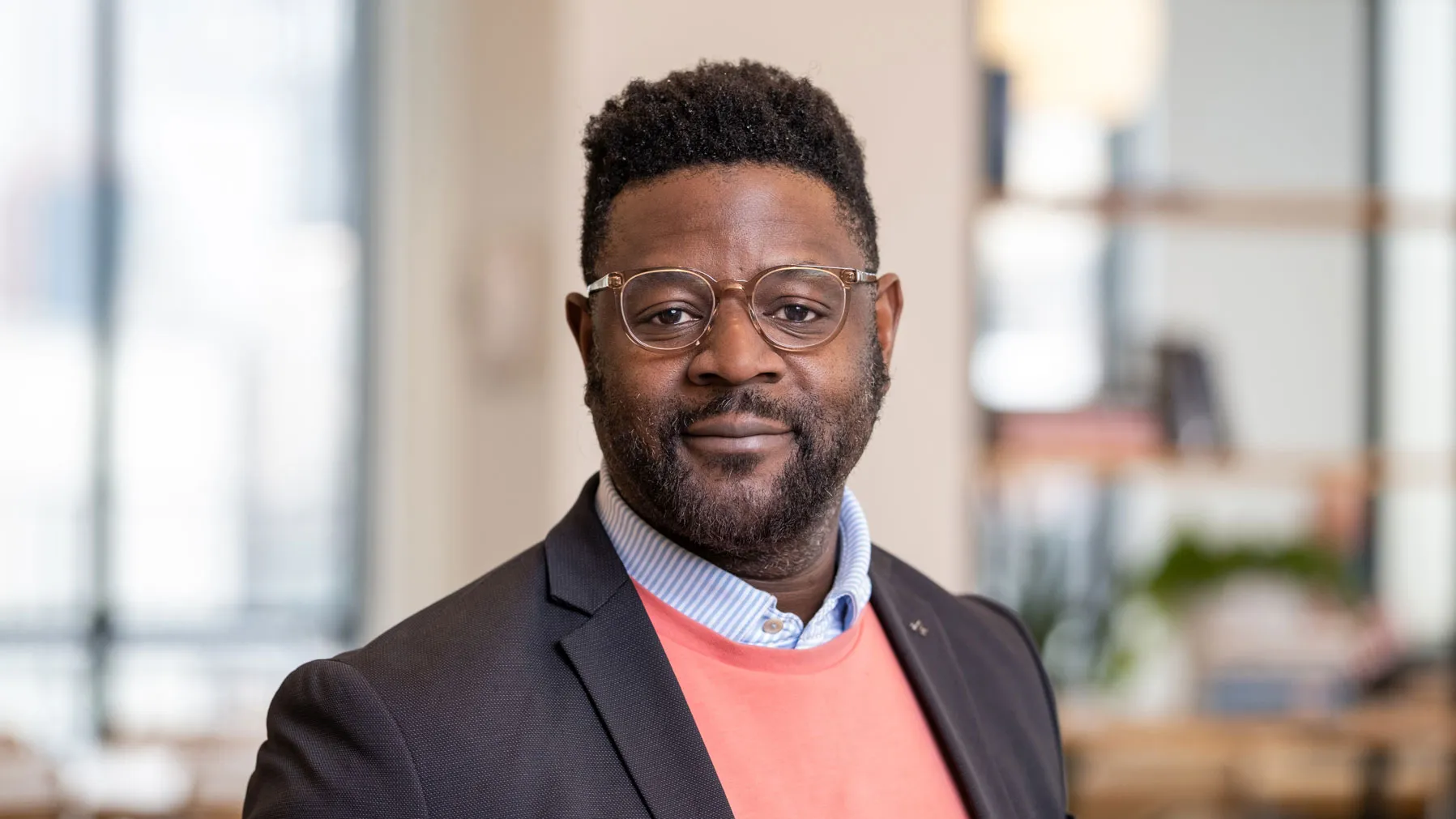 A Black man slightly smiles as he poses for this portrait in a modern office setting with shelves and large windows in the background. He wears glasses and sport coat over over a collared shirt and sweater.