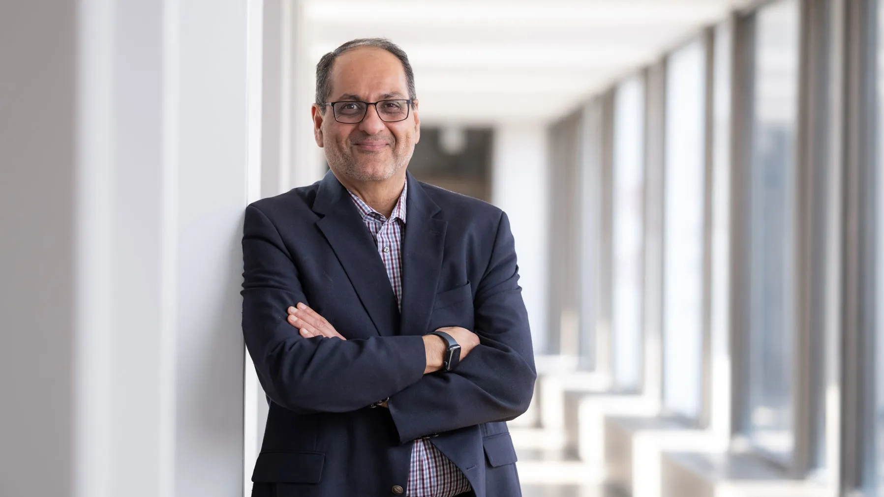 Ness Shroff, a man of Indian descent, smiles as he leans against a wall in a bright hallway at Ohio State. He has arms crossed and wears glasses, a suit jacket and patterned shirt. He looks like someone who would be fascinating to have a discussion with. Large windows line the corridor in the background.