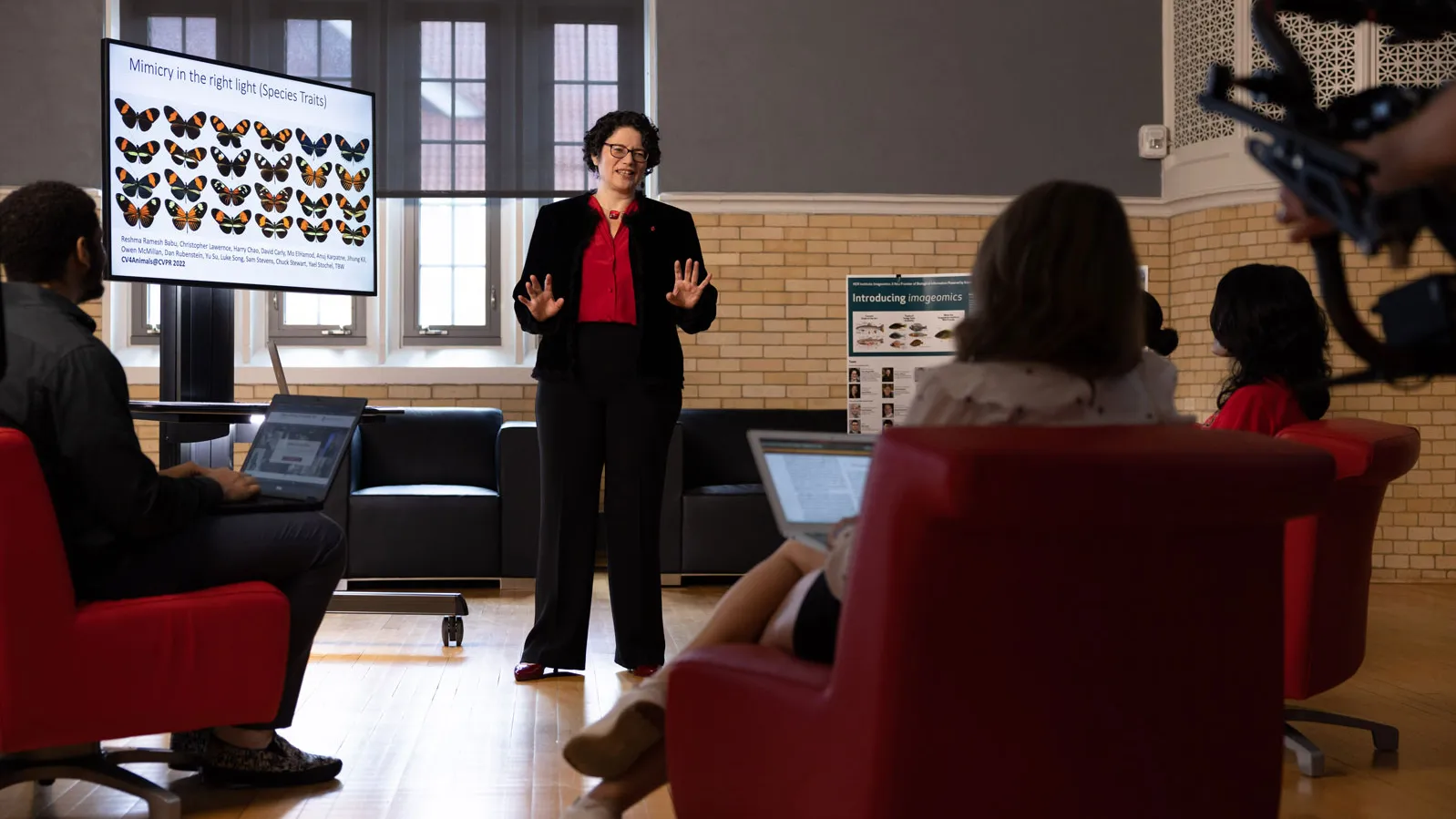 Tanya Berger Wolf, a white woman with short curly hair and. pants suit, stands at the front of a small room giving a presentation while several people seated in chairs watch and use laptops. A large screen beside the presenter displays images of butterflies with labels. The setting appears to be an indoor meeting or classroom space with tall windows and light-colored walls.
