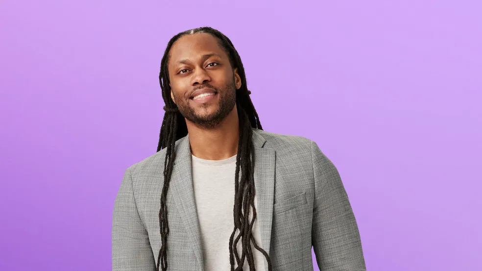Victor St. John, a black man with long dreadlock hair, wearing a blazer and jeans, looks friendly and studious—like he'd be the coolest professor—as he poses for this photo.  