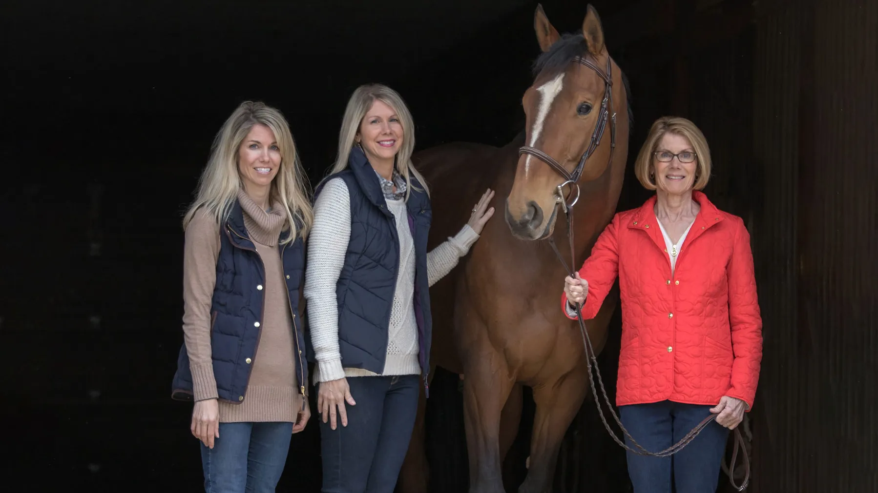 Three people stand beside a horse inside a barn entrance, with the mom holding the horse’s lead rope and her two grown daughters standing on the other side of the horse. They all smile as they pose.