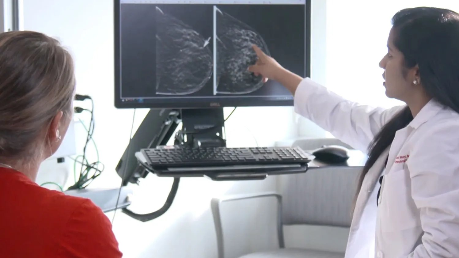A medical professional in a white coat points to breast imaging scans on a computer monitor while another person sits nearby, looking at the screen.